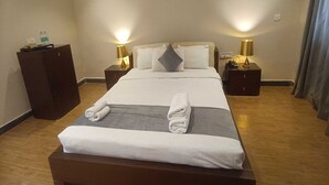 Een net opgemaakt bed met witte lakens en handdoeken, twee nachtkastjes met lampen, een houten kledingkast en een effen muur.
