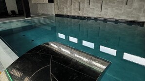 Una piscina interior con agua cristalina, una plataforma de salto negra e iluminación verde en el techo.