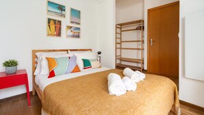 Ein ordentlich bezogenes Bett mit bunten Kissen, ein roter Nachttisch mit Pflanze, eine Holzleiter-Regal und eine Holztür.