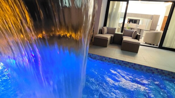 Piscina interna com cascata, área de estar com sofás e cozinha visível através de uma porta de vidro.