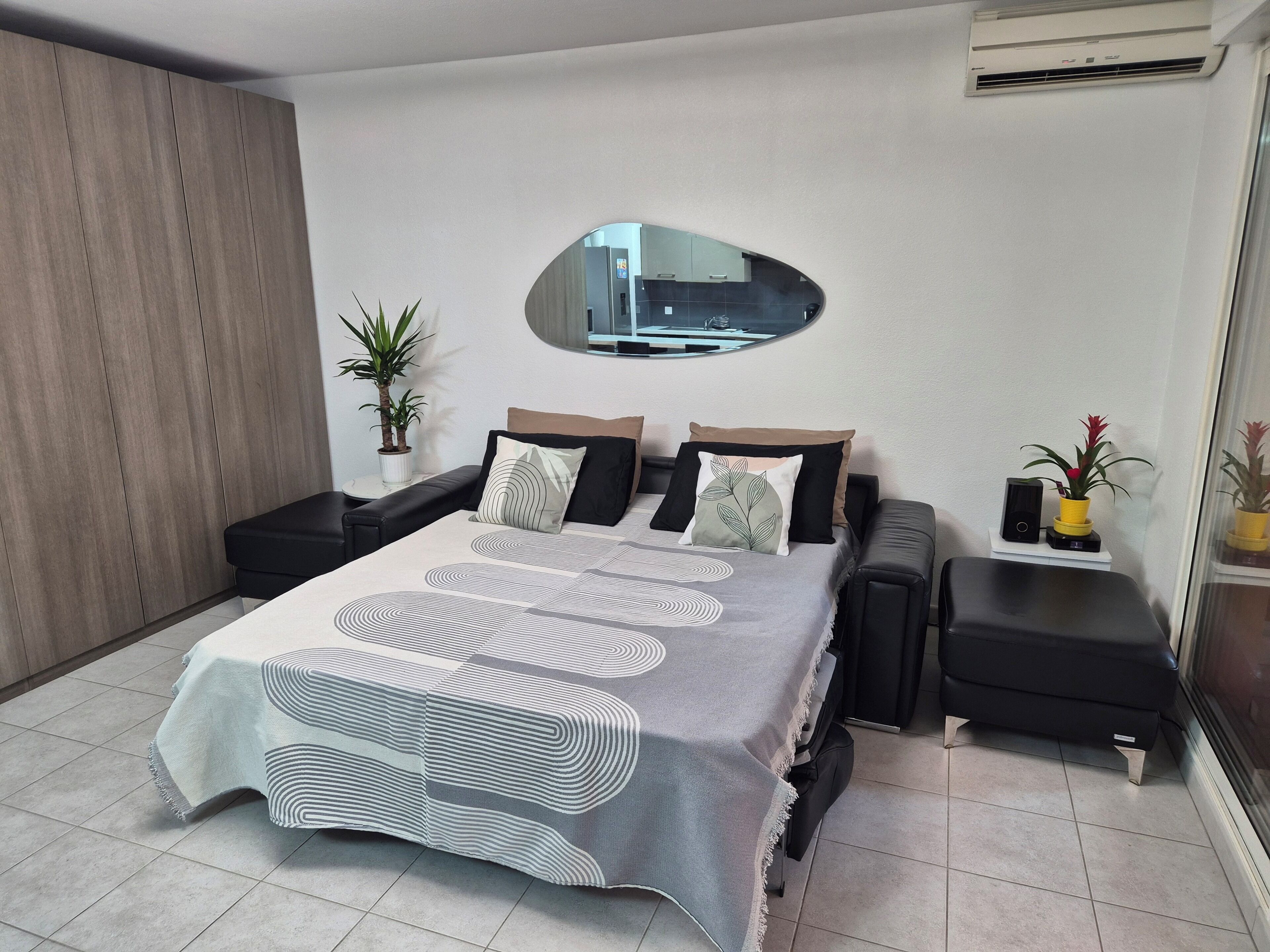 A modern hotel room with a large bed, a small side table, a potted plant, and a mirror on the wall.