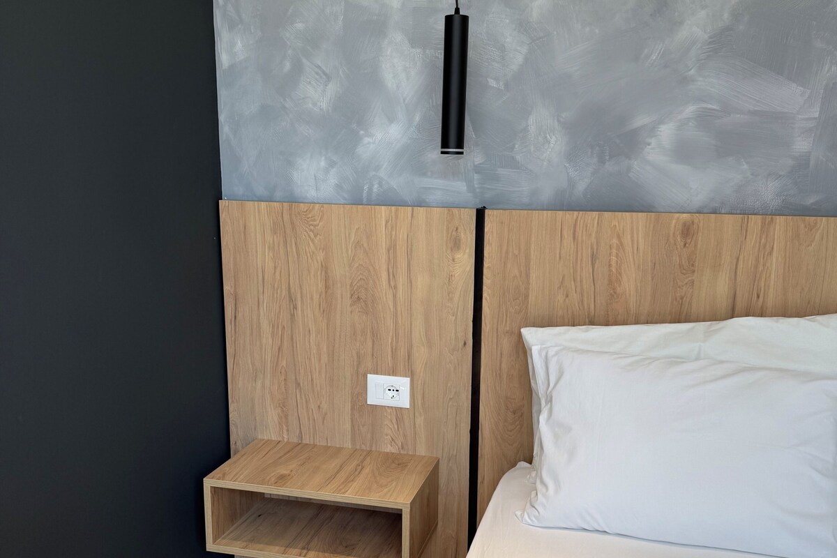 Een moderne slaapkamer met een houten hoofdbord, nachtkastje, een wit bed en een gestructureerde grijze muur.