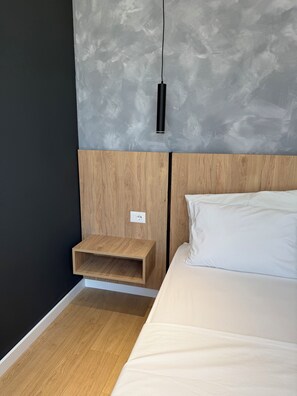 Een moderne slaapkamer met een houten hoofdbord, nachtkastje, een wit bed en een gestructureerde grijze muur.