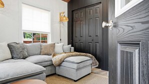 Ein modernes Wohnzimmer mit einem Ecksofa, einem kleinen Hocker, einem Korbtisch, einer dunkelholzigen Tür und einem Fenster mit Jalousien.
