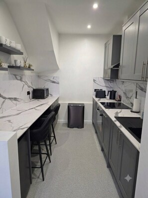 Uma cozinha compacta com bancada de mármore, banquetas, armários cinzas e uma lixeira preta.