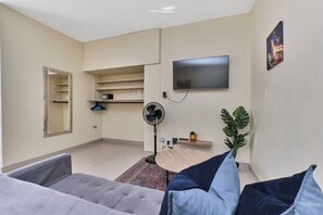 A living room with a grey sofa, a standing fan, a flat-screen TV, a mirror, and a plant. - Urban Luxe Studio Illovo Mews by ctha (Sandton)