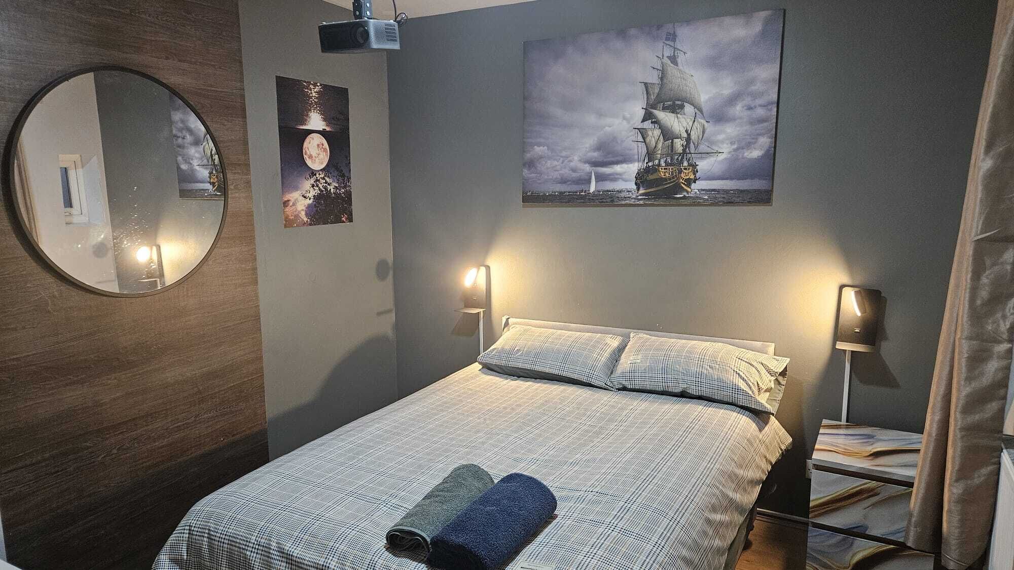 A bedroom with a bed, a round mirror, wall-mounted lamps, and a framed picture of a ship.