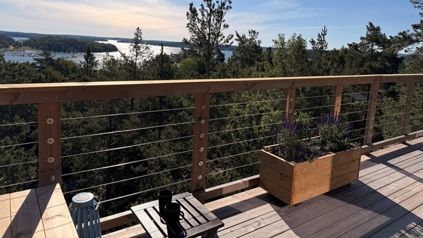 Una terraza de madera con barandilla, un banco, dos tazas negras y una jardinera con plantas.