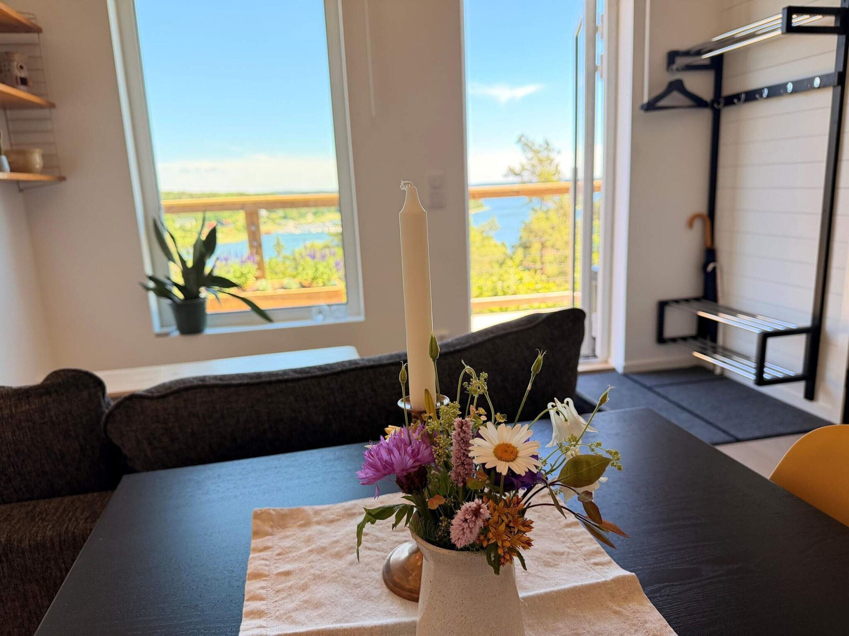 Una habitación con una mesa de comedor, un florero con flores, una vela, una ventana que ofrece vistas a la vegetación y un puente, además de un estante de metal negro.