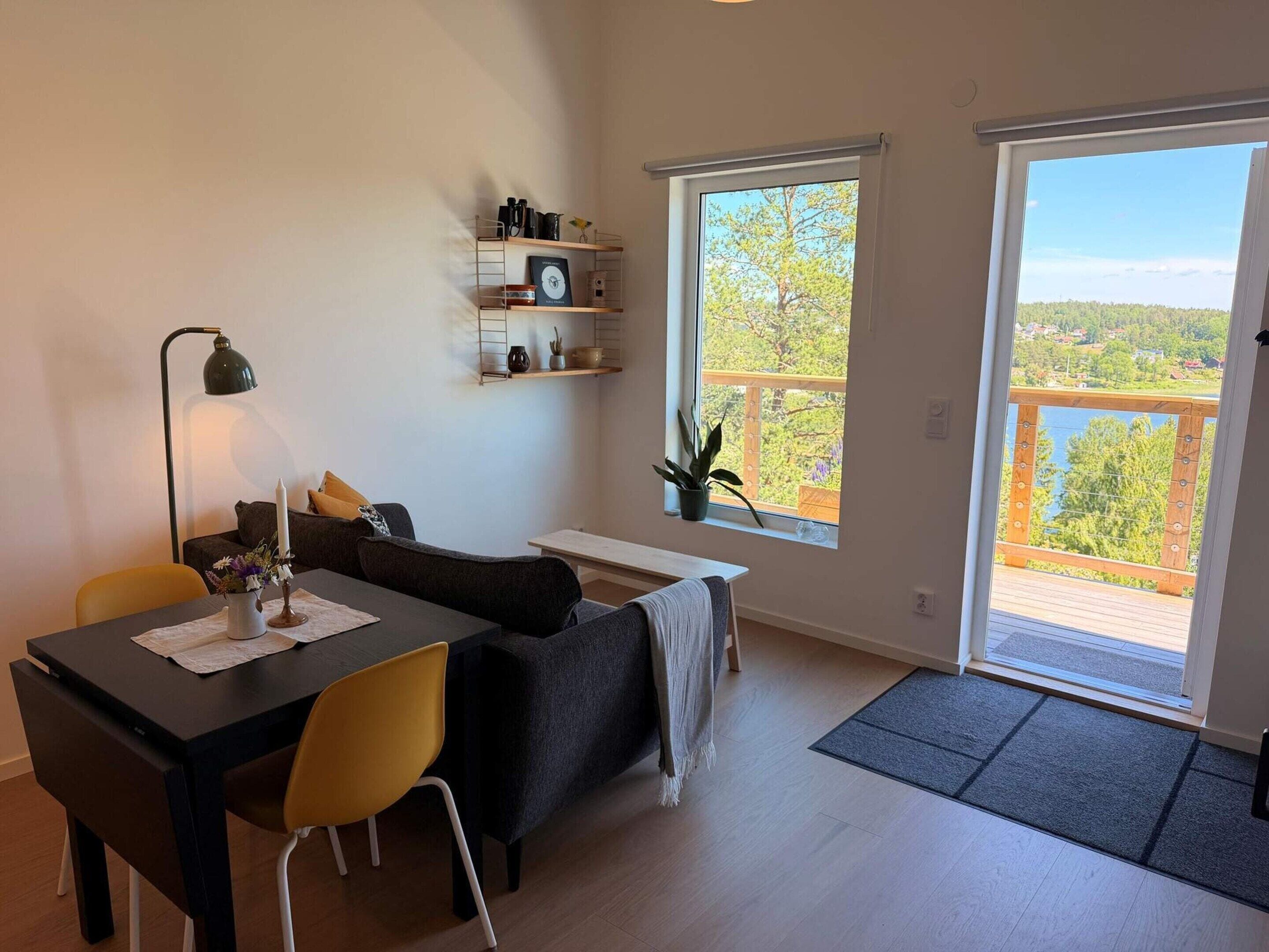 Un espacio habitable moderno con comedor, sofá, una mesita, lámpara, planta y un balcón con vistas.