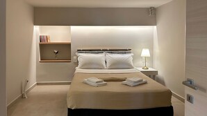 A modern hotel room with a neatly made bed, a bedside table with a lamp, a shelf with books, and a small vase. - Casa Verde | Minimal & Cozy Stay (Elliniko)