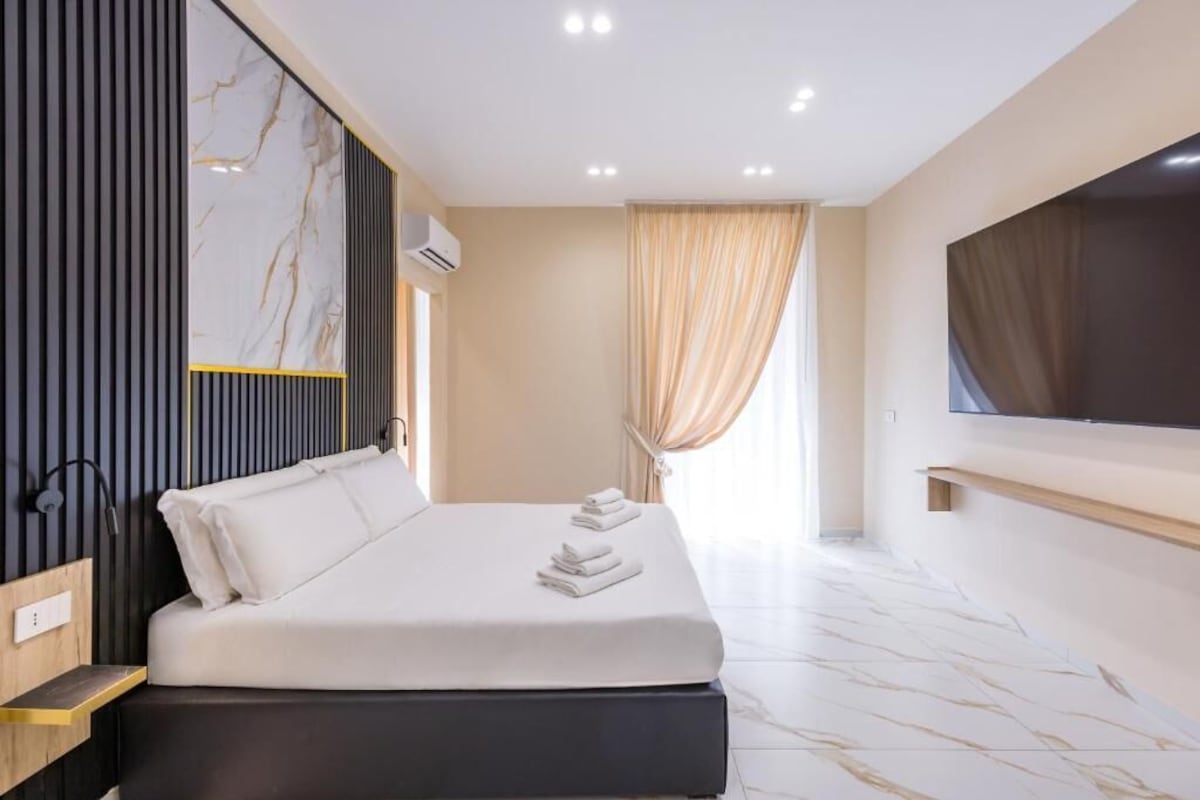 Una moderna camera d'hotel con un letto grande, una televisione a schermo piatto, una parete con motivo marmorizzato e una finestra con tende.