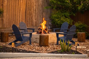 A fire pit with logs, surrounded by Adirondack chairs, set against a wooden fence.