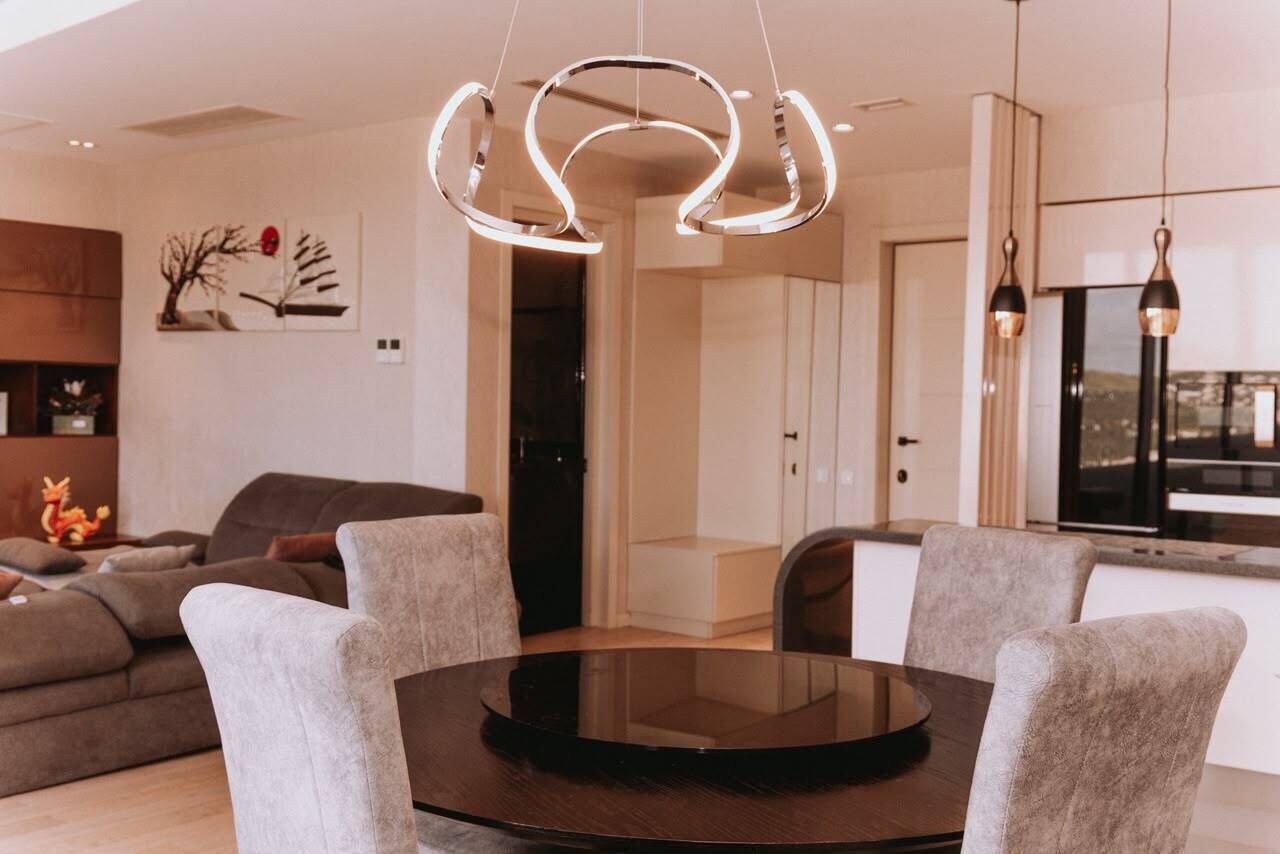 A modern living room with a round glass table, grey upholstered chairs, a contemporary light fixture, and a minimalist decor.