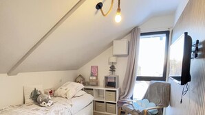 Een slaapkamer met een schuine kap, een bed met een witte dekbedovertrek, een stoel, een bureau met opbergruimte, een lamp en een tv aan de muur.
