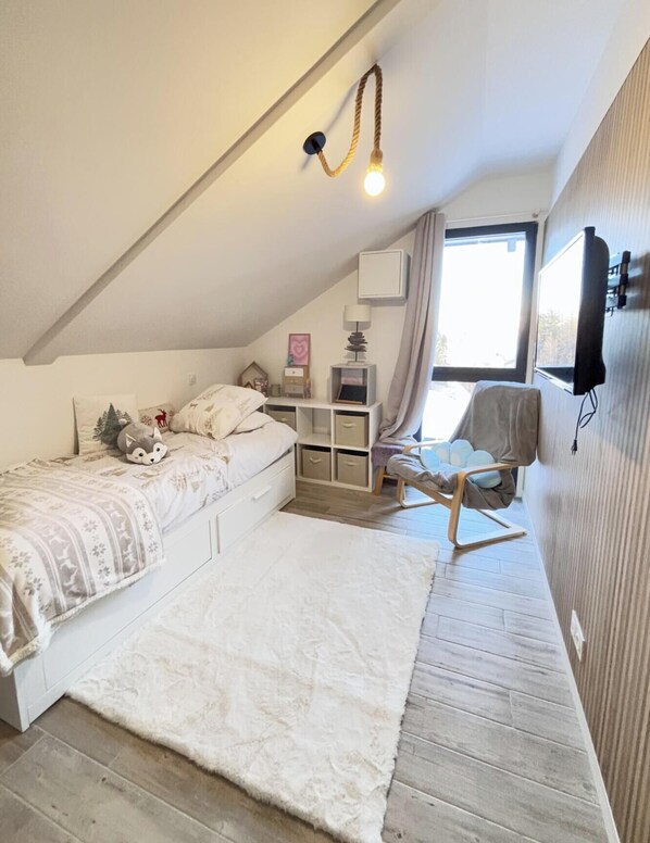Une chambre avec un plafond en pente, un lit recouvert d’une couette blanche, une chaise, un bureau avec des rangements, une lampe et un téléviseur fixé au mur.