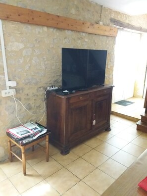 A room with a television on a wooden cabinet, a small table with magazines, and a stone wall. - La Grange Vacation Rental in the Périgord Noir (Allas-les-Mines)