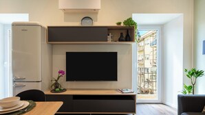 Ein modernes Wohnzimmer mit Flachbildfernseher, integrierter Klimaanlage, Essbereich mit Tisch und Stühlen und Blick auf ein Mehrfamilienhaus durch ein großes Fenster.