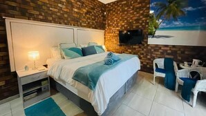 Un dormitorio con pared de ladrillo, una cama con ropa blanca, una mesita de noche, un televisor y un cuadro de una playa.