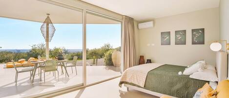 A bedroom with a bed, a dining area with a table and chairs, and a view through large windows.