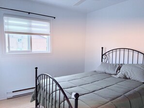 Ein Einzelbett mit Metallgestell und einer grauen Tagesdecke, ein Fenster mit Jalousien und eine weiße Wand.