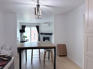 A modern kitchen with a wooden table, bar stools, a ceiling light fixture, and a view of a red brick building through the window. - Cozy 2-bedroom apartment 22 minutes from Montreal (La Prairie)
