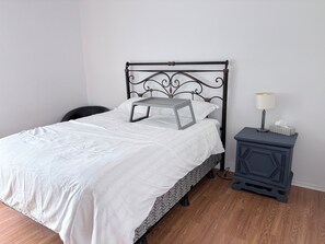 A neatly made bed with a white bedspread and a gray tray, a bedside table with a lamp and tissues, and a chair in a room with wooden flooring. - Cozy 2-bedroom apartment 22 minutes from Montreal (La Prairie)