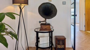 Una stanza con una lampada da terra, un grammofono, un tavolino con un dispositivo bianco e un armadietto in legno.