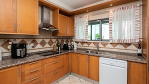A kitchen with wooden cabinets, a dishwasher, a coffee maker, and a window with striped curtains. - Moradia V3 a 100m da praia by ALGARVEMANTA (24V3) (Manta Rota)