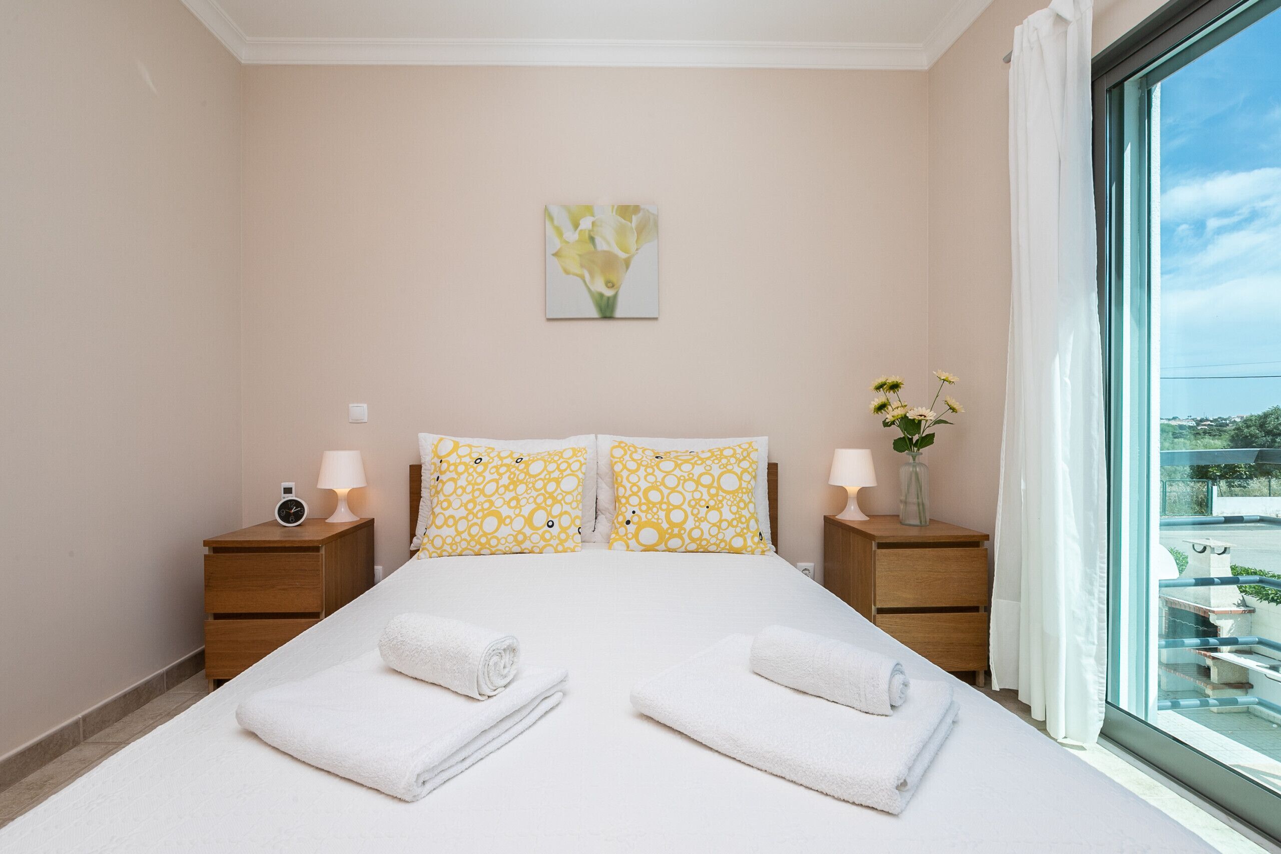 A bedroom with a bed, two bedside tables, a nightstand, a window with a view, and a painting on the wall.