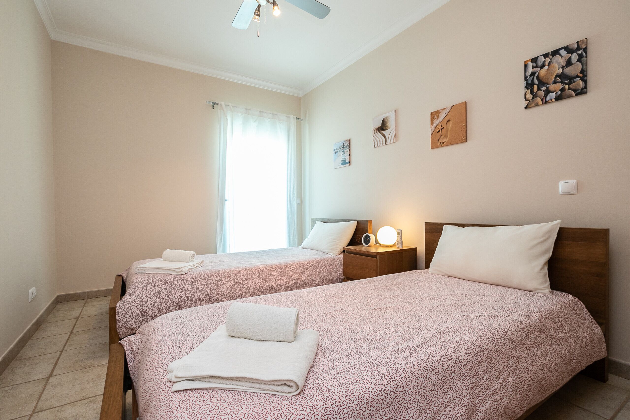 Hotellihuone, jossa on kaksi sänkyä, kattotuuletin, yöpöytä lampun kanssa ja taideteoksia seinillä.