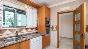 A kitchen with a dishwasher, oven, microwave, and a window with curtains. - Moradia V3 a 100m da praia by ALGARVEMANTA (24V3) (Manta Rota)