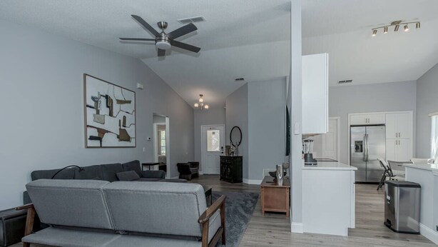 Una sala moderna con un sofá gris, un ventilador de techo, una zona de cocina con armarios blancos y una mesa de comedor.