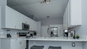 Cocina moderna con armarios blancos, una isla central con taburetes, electrodomésticos de acero inoxidable y una lámpara colgante.