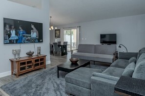 Ein modernes Wohnzimmer mit einer grauen Couch, einem Flachbildfernseher, einer Holzkabine und einem Essbereich mit Tisch und Stühlen.