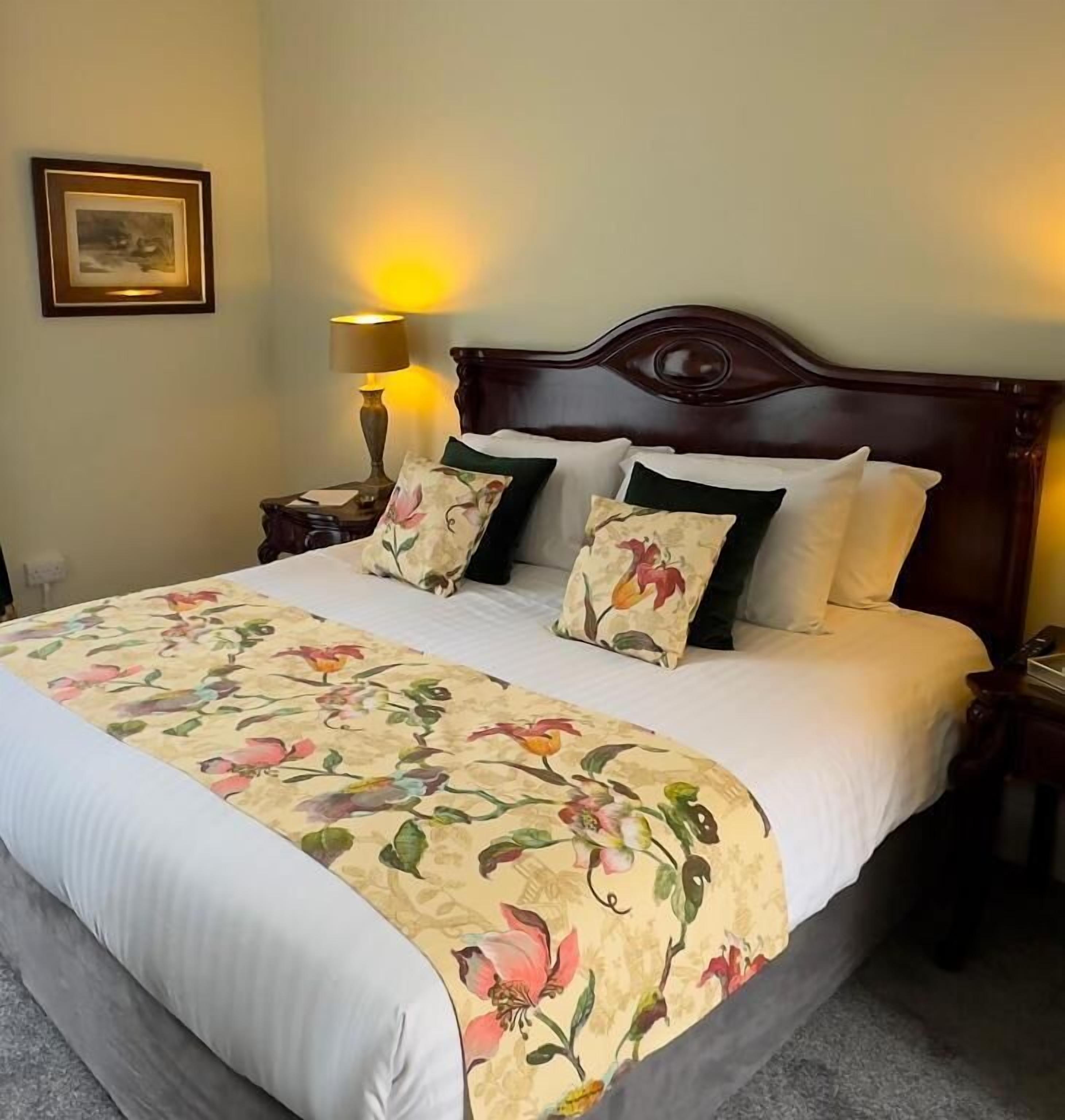 Un lit recouvert d’une couvre-lit à motifs floraux, deux oreillers, une table de chevet avec une lampe et un tableau encadré accroché au mur.