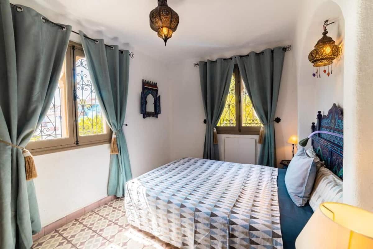 Un dormitorio con cama, cortinas, una lámpara de pared y una ventana con celosía decorativa de metal.