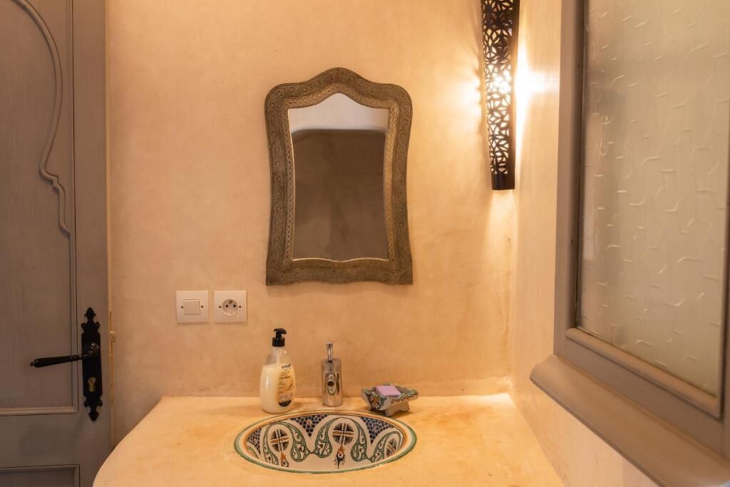 A bathroom with a beige wall, a mirror, a sink with a decorative basin, and a wall-mounted light fixture.