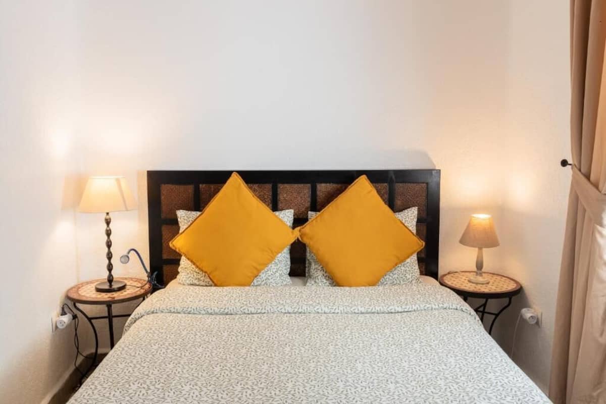 Una cama con una colcha estampada, dos almohadas iguales y dos lámparas de noche a cada lado.