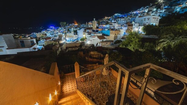 A nighttime view of a hillside town with buildings illuminated, a staircase with ornate railings, and a clear sky.