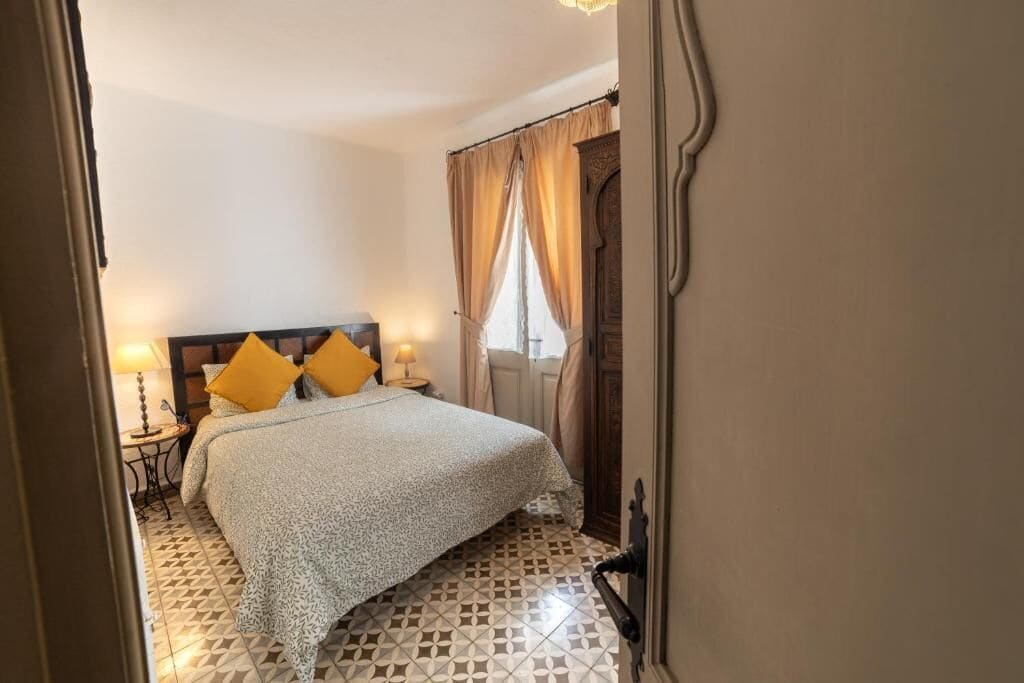 A bedroom with a bed, two bedside tables, a patterned floor, and a window with curtains.
