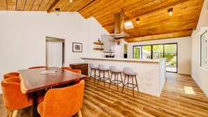 A modern interior with a wooden ceiling, a dining area with orange chairs, a kitchen with bar stools, and a large window.