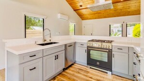 A kitchen with a stove, sink, and cabinets, wooden ceiling, and windows.
