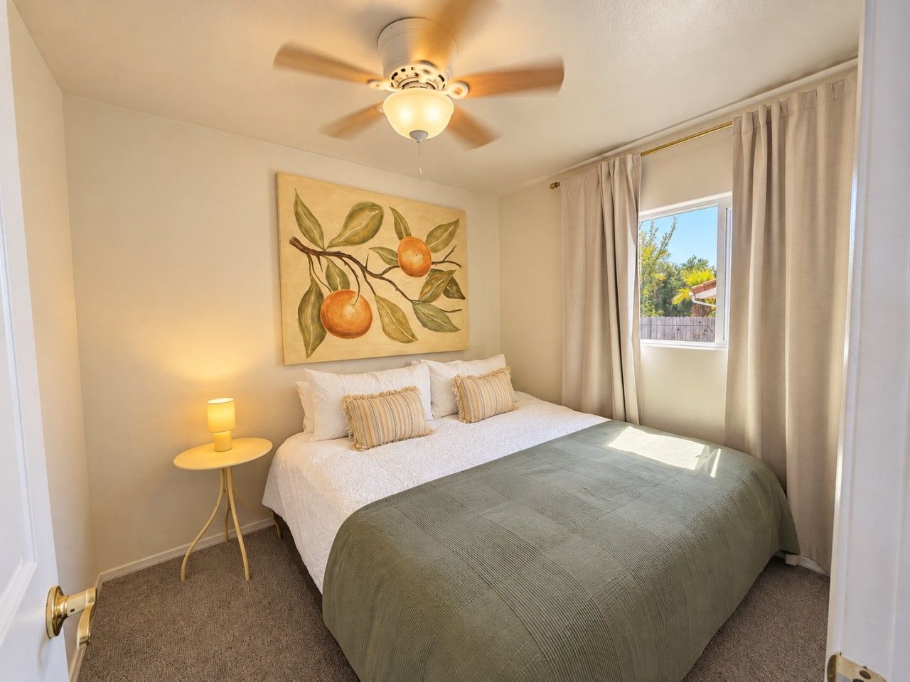 A bedroom with a bed, a nightstand, a ceiling fan, a window with curtains, and a wall painting.