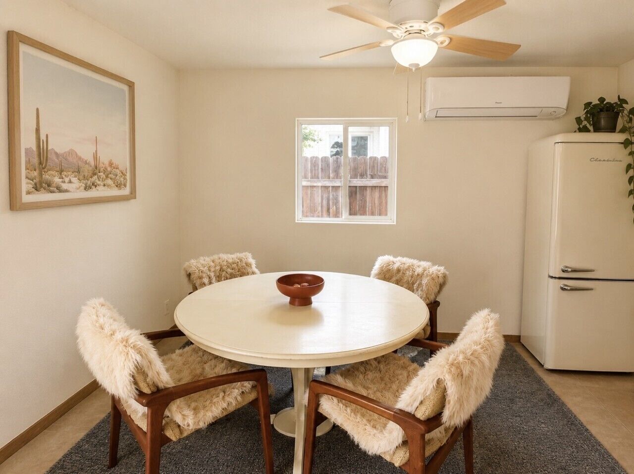 A dining area with a round table, four chairs, a ceiling fan, a window, and a refrigerator.