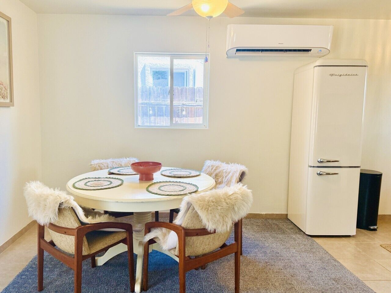 A dining area with a round table, chairs with fur throw pillows, a red bowl, and a white refrigerator.