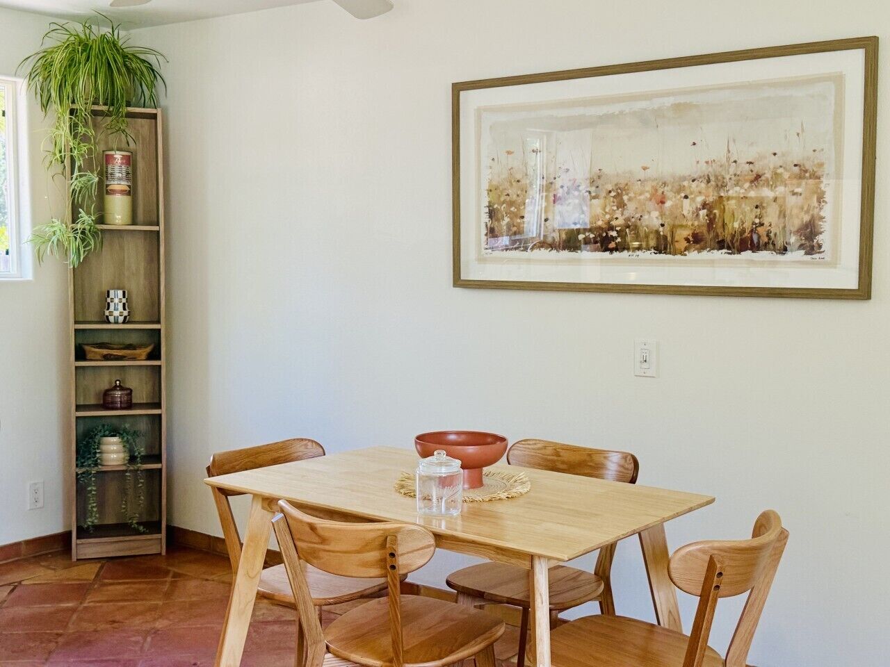 A dining area with a wooden table and chairs, a red bowl, and a framed artwork on the wall.