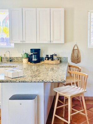A kitchen with white cabinets, a granite countertop, a coffee maker, a basket, and a bag hanging on the wall. - Charming 2BR Ojai Escape (Ojai)