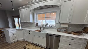 A kitchen with white cabinetry, a stainless steel dishwasher, a farmhouse sign, and a window with blinds.