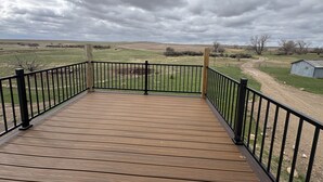 A wooden deck with black railings, overlooking a grassy field and a dirt path, with a cloudy sky above.